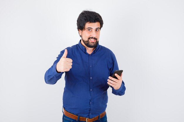 Varón joven que muestra el pulgar hacia arriba mientras sostiene el teléfono en la camisa azul real y parece contento. vista frontal.