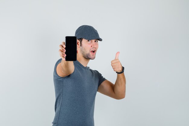 Varón joven que muestra el pulgar hacia arriba mientras sostiene el teléfono móvil en la gorra de la camiseta