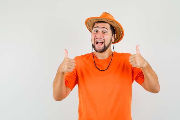 Varón joven que muestra los pulgares dobles para arriba en la camiseta anaranjada, el sombrero y parece afortunado. vista frontal.