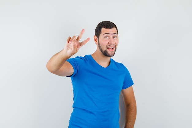 Varón joven que muestra el signo de la victoria en la camiseta azul y mirando alegre