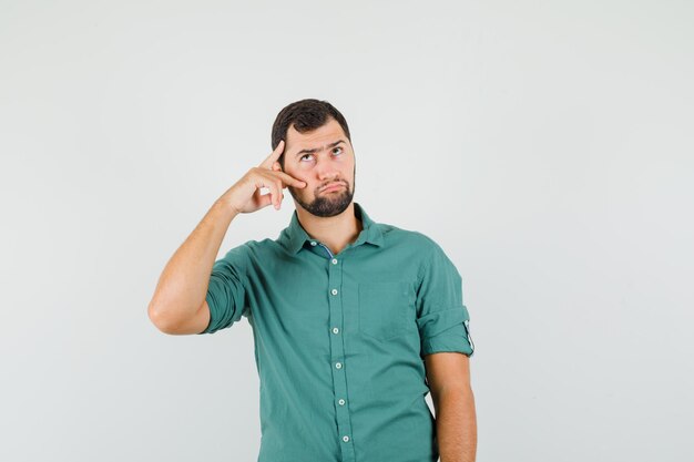 Varón joven que muestra v-sign en su rostro con camisa verde y mirando pensativo, vista frontal.