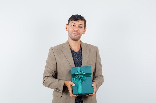 Varón joven que sostiene la caja de regalo azul en la chaqueta marrón grisácea y que parece contento. vista frontal.