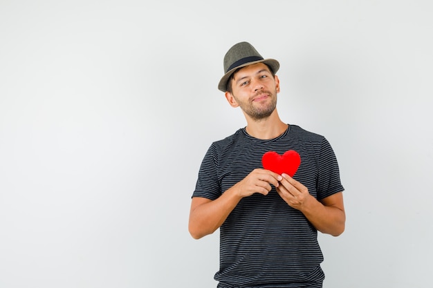 Foto gratuita varón joven que sostiene el corazón rojo en el sombrero de la camiseta y que parece alegre