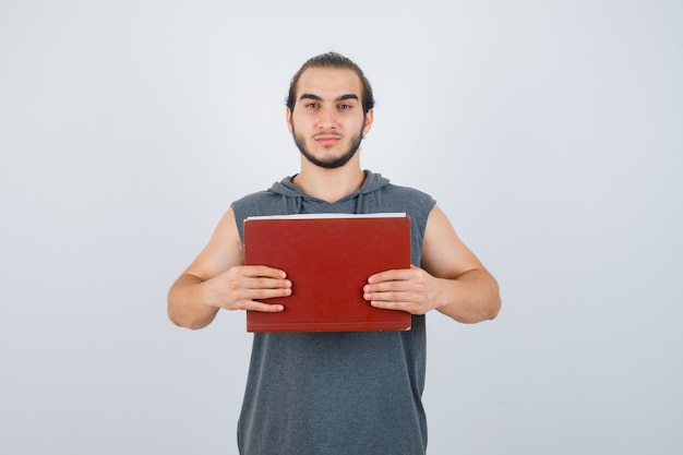 Varón joven que sostiene el libro delante de él en una sudadera con capucha sin mangas y que parece serio. vista frontal.