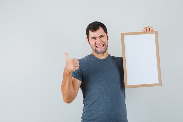Varón joven que sostiene el marco vacío que muestra el pulgar hacia arriba en la camiseta gris y que parece alegre