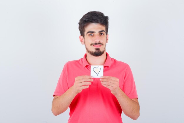 Varón joven que sostiene una nota adhesiva en la camiseta y que mira linda, vista frontal.