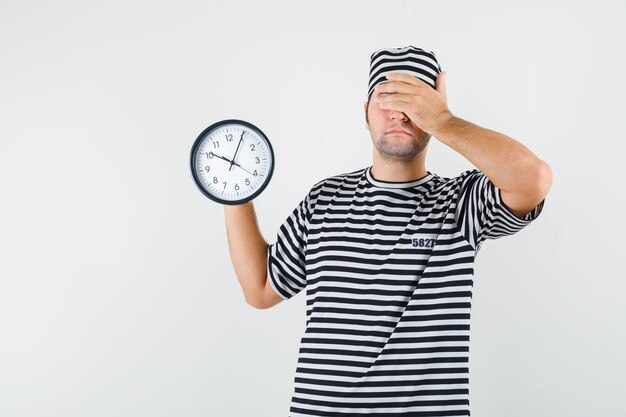 Varón joven que sostiene el reloj de pared en camiseta, sombrero y mirando olvidadizo, vista frontal.