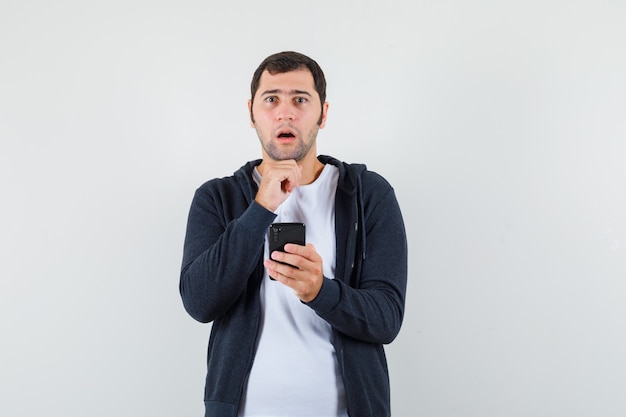 Varón joven que sostiene el teléfono móvil en la camiseta, la chaqueta y que mira desconcertado. vista frontal.