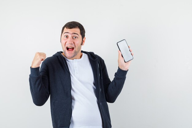 Varón joven que sostiene el teléfono móvil en camiseta, chaqueta y que parece feliz. vista frontal.