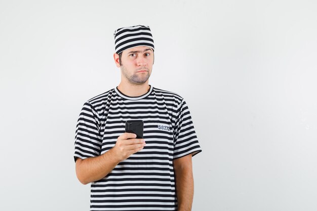 Varón joven que sostiene el teléfono móvil en la camiseta, sombrero y que mira vacilante. vista frontal.