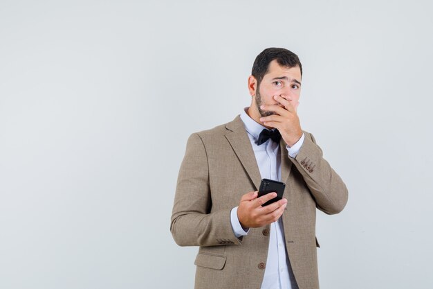 Varón joven que sostiene el teléfono móvil en traje y parece olvidadizo. vista frontal.