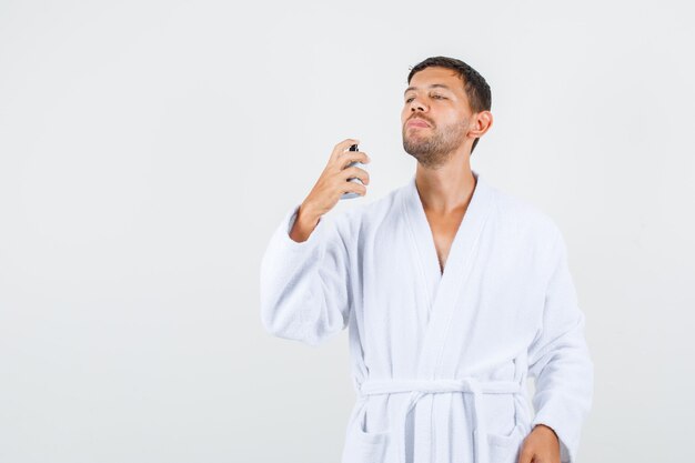 Varón joven rociando perfume en bata de baño blanca y mirando confiado, vista frontal.