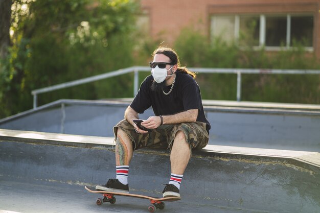 Varón joven sentado con una patineta en el parque con una mascarilla médica - Concepto Covid-19