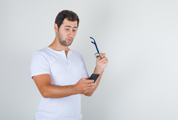 Varón joven con smartphone y sosteniendo gafas en camiseta blanca y mirando sorprendido