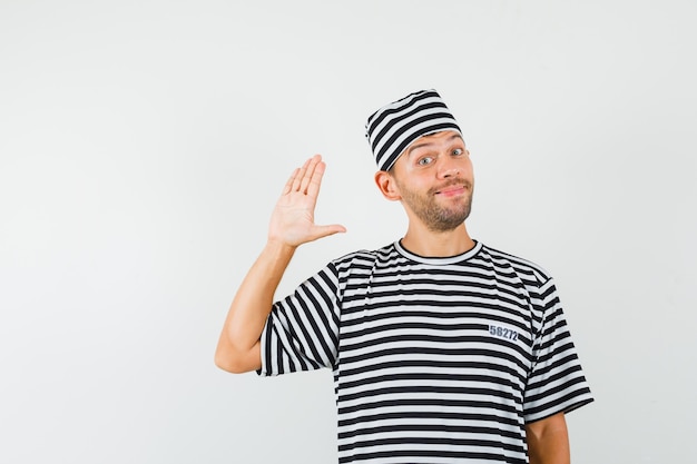 Varón joven con sombrero de camiseta a rayas agitando la mano para saludar y mirar alegre