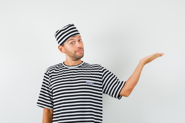 Varón joven con sombrero de camiseta a rayas extendiendo la palma a un lado y mirando confiado