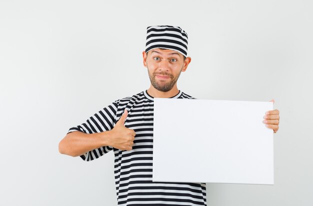 Varón joven con sombrero de camiseta a rayas sosteniendo un lienzo en blanco mostrando el pulgar hacia arriba y mirando feliz