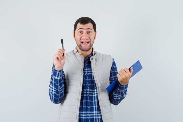 Varón joven sonriendo mientras sostiene la pluma y el cuaderno en camisa, chaqueta y mirando contento, vista frontal.