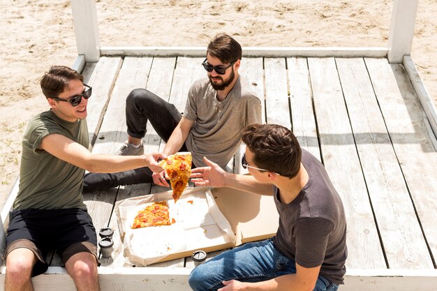 Varón joven sonriente que da la rebanada de amigo de la pizza en la playa