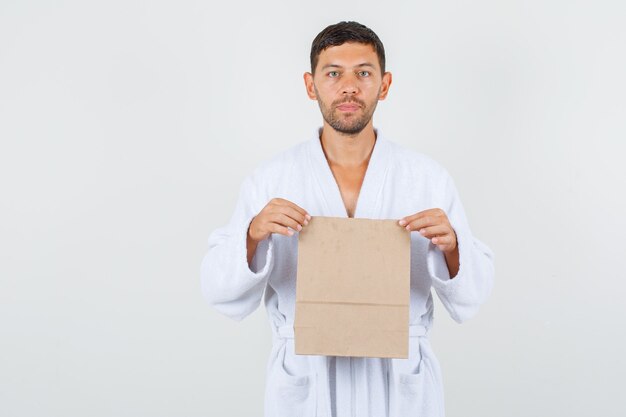 Varón joven sosteniendo una bolsa de papel artesanal en bata de baño blanca y mirando estricta, vista frontal.