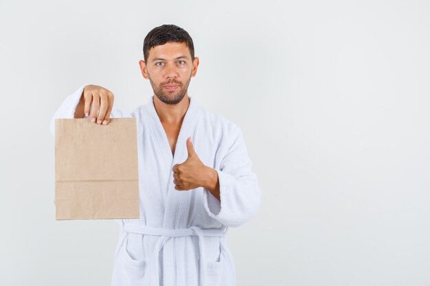 Varón joven sosteniendo la bolsa de papel con el pulgar hacia arriba en bata de baño blanca y mirando complacido, vista frontal.