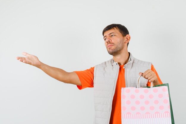 Varón joven sosteniendo bolsas de la compra, estirando la mano en camiseta, chaqueta, vista frontal.