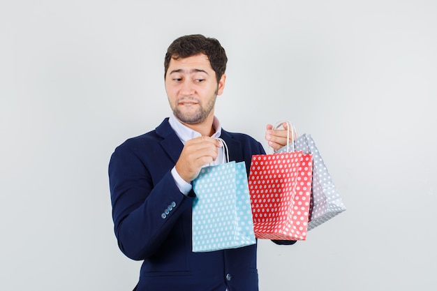 Varón joven sosteniendo bolsas de papel en traje azul y mirando curioso, vista frontal.