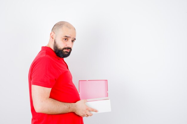 Varón joven sosteniendo caja de regalo abierta en camiseta roja y mirando pensativo, vista frontal.