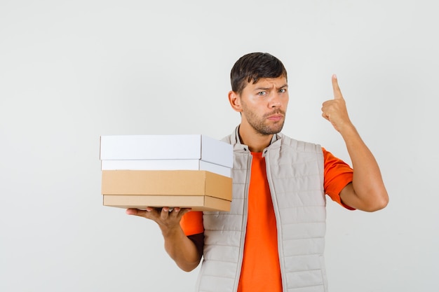 Varón joven sosteniendo cajas de cartón, apuntando hacia arriba en camiseta, chaqueta, vista frontal.