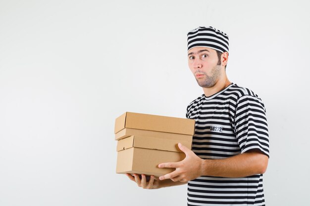 Varón joven sosteniendo cajas de cartón en camiseta, sombrero y mirando sorprendido. vista frontal.