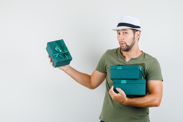 Varón joven sosteniendo cajas presentes en camiseta verde y sombrero y mirando pensativo