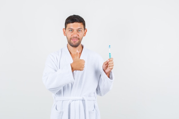 Varón joven sosteniendo el cepillo de dientes con el pulgar hacia arriba en bata de baño blanca y mirando positivo, vista frontal.