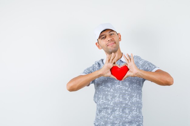 Varón joven sosteniendo corazón rojo en camiseta y gorra y mirando alegre