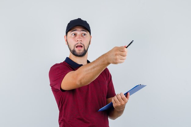 Varón joven sosteniendo cuaderno y bolígrafo en camiseta roja, gorra negra y mirando con cuidado, vista frontal.