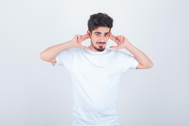 Varón joven sosteniendo los dedos detrás de las orejas en camiseta y mirando divertido, vista frontal.