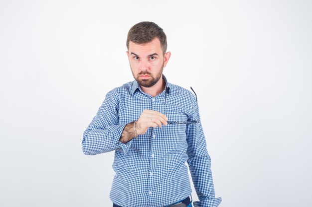 Varón joven sosteniendo gafas mientras mira a la cámara en camisa, jeans y se ve guapo. vista frontal.