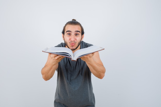 Varón joven sosteniendo un libro abierto en una sudadera con capucha sin mangas y mirando confiado, vista frontal.