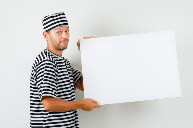 Varón joven sosteniendo un lienzo en blanco con un sombrero de camiseta a rayas y mirando alegre