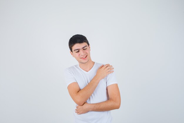 Varón joven sosteniendo la mano en el hombro en camiseta y mirando alegre, vista frontal.