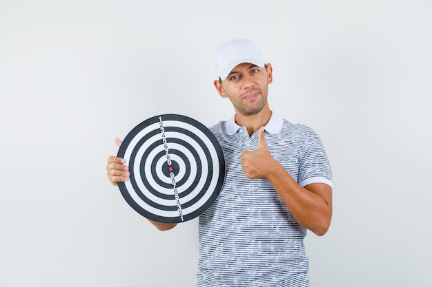 Foto gratuita varón joven sosteniendo el tablero de dardos con el pulgar hacia arriba en camiseta y gorra y mirando complacido