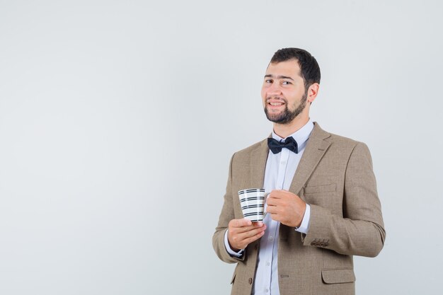 Varón joven sosteniendo una taza de bebida en traje y mirando alegre, vista frontal.