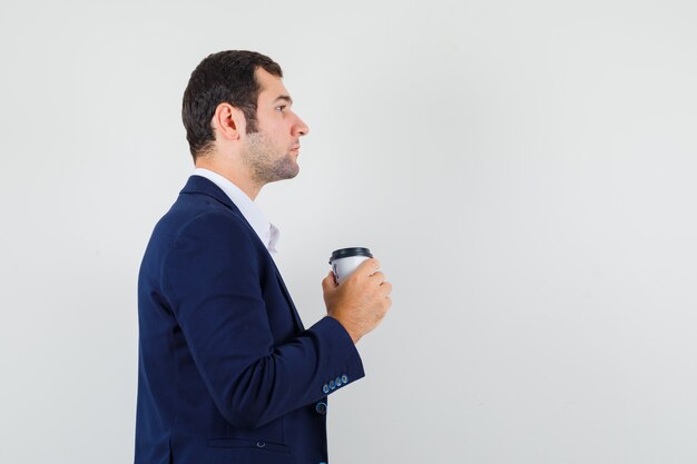 Varón joven sosteniendo una taza de café en camisa, chaqueta y mirando enfocado