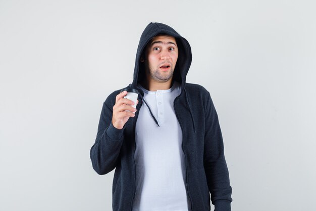Varón joven sosteniendo una taza de café en camiseta, chaqueta y mirando desconcertado. vista frontal.
