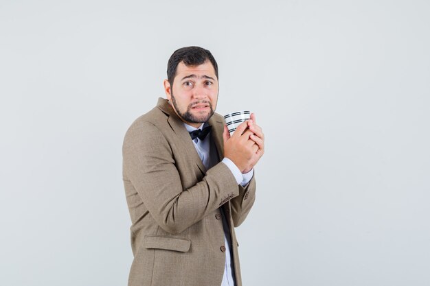 Varón joven sosteniendo una taza de café en traje y mirando humilde, vista frontal.