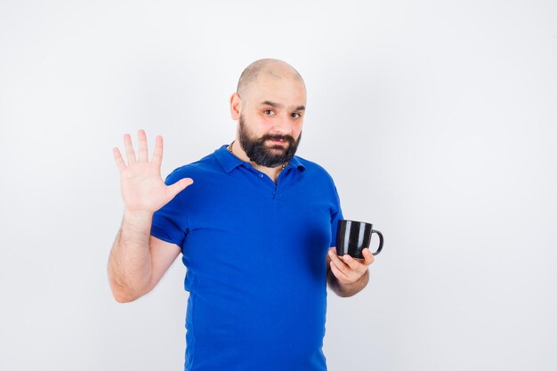 Varón joven sosteniendo la taza mientras agita la mano para saludar en camisa azul, vista frontal.