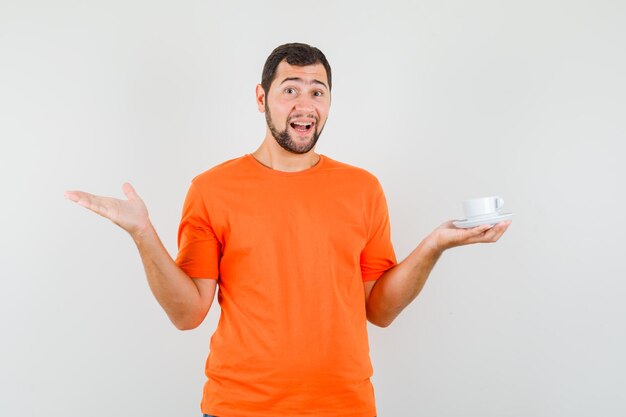 Varón joven sosteniendo la taza con platillo en camiseta naranja y mirando jovial. vista frontal.