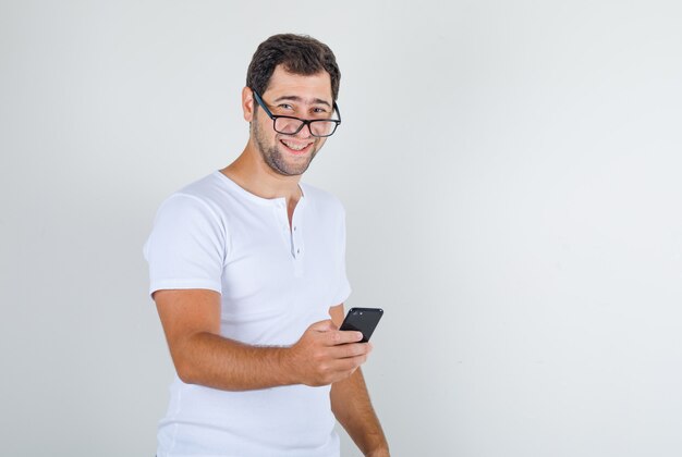 Varón joven sosteniendo teléfono inteligente y riendo con camiseta blanca, gafas y mirando feliz