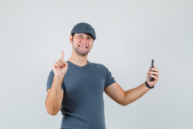 Varón joven sosteniendo el teléfono móvil apuntando hacia arriba en la gorra de la camiseta y mirando feliz
