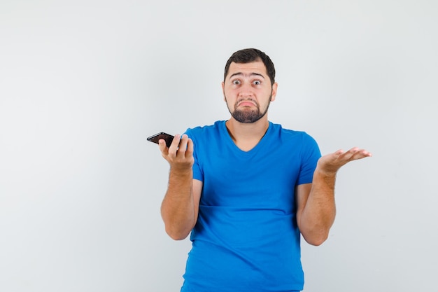 Varón joven sosteniendo teléfono móvil en camiseta azul y mirando confundido