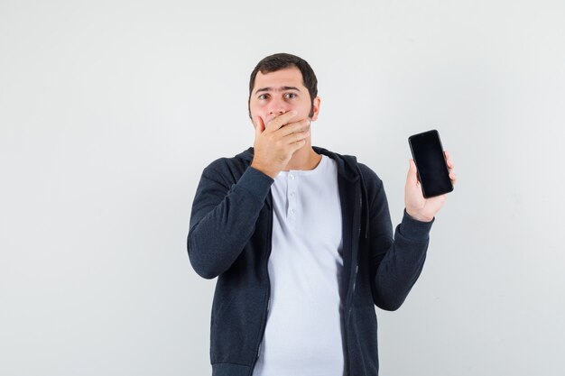 Varón joven sosteniendo teléfono móvil en camiseta, chaqueta y mirando sorprendido. vista frontal.